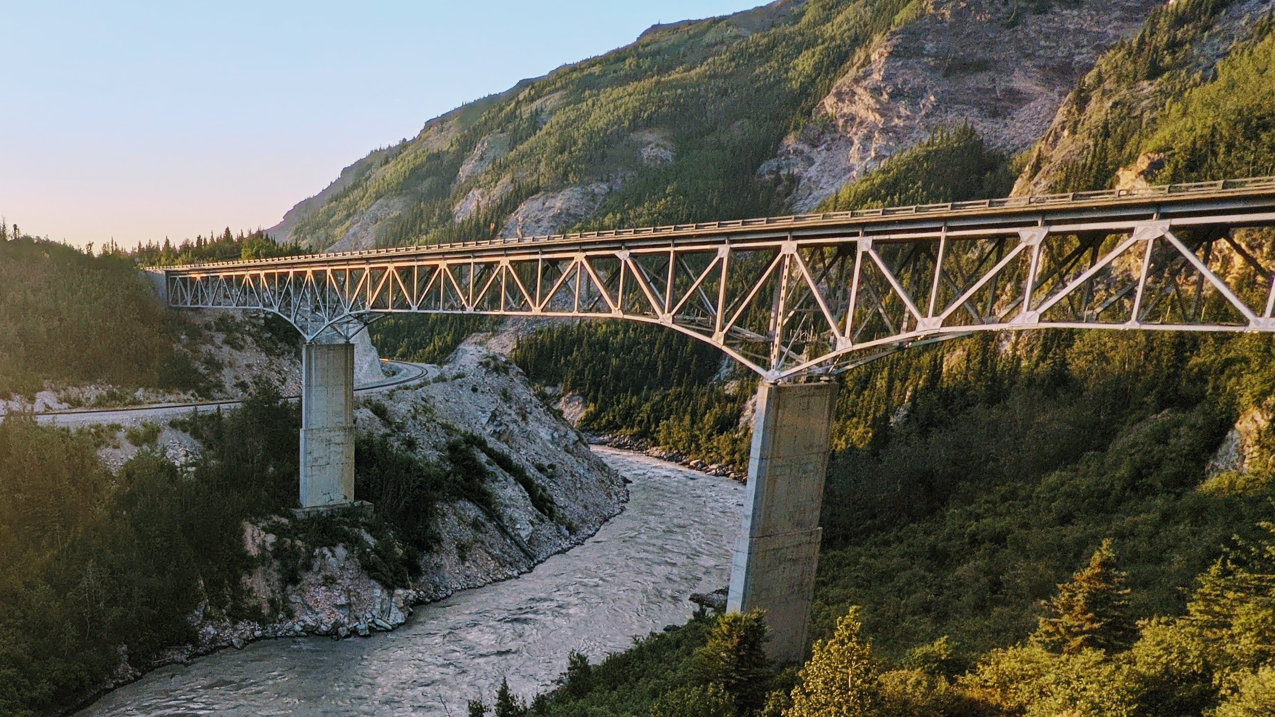 Picture of a bridge in the Alaskan wilderness over a rushing river. The weather is clear and it's apparent that it's a 'golden hour' photo.